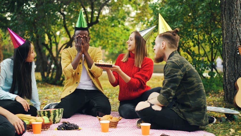 Friends in party hats celebrating a birthday with cake on a picnic blanket