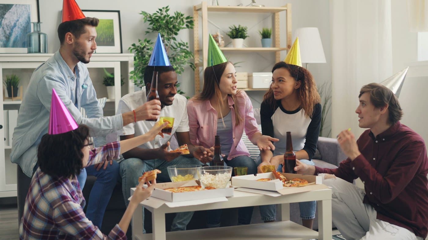 Guests in party hats sharing pizza and drinks around a coffee table